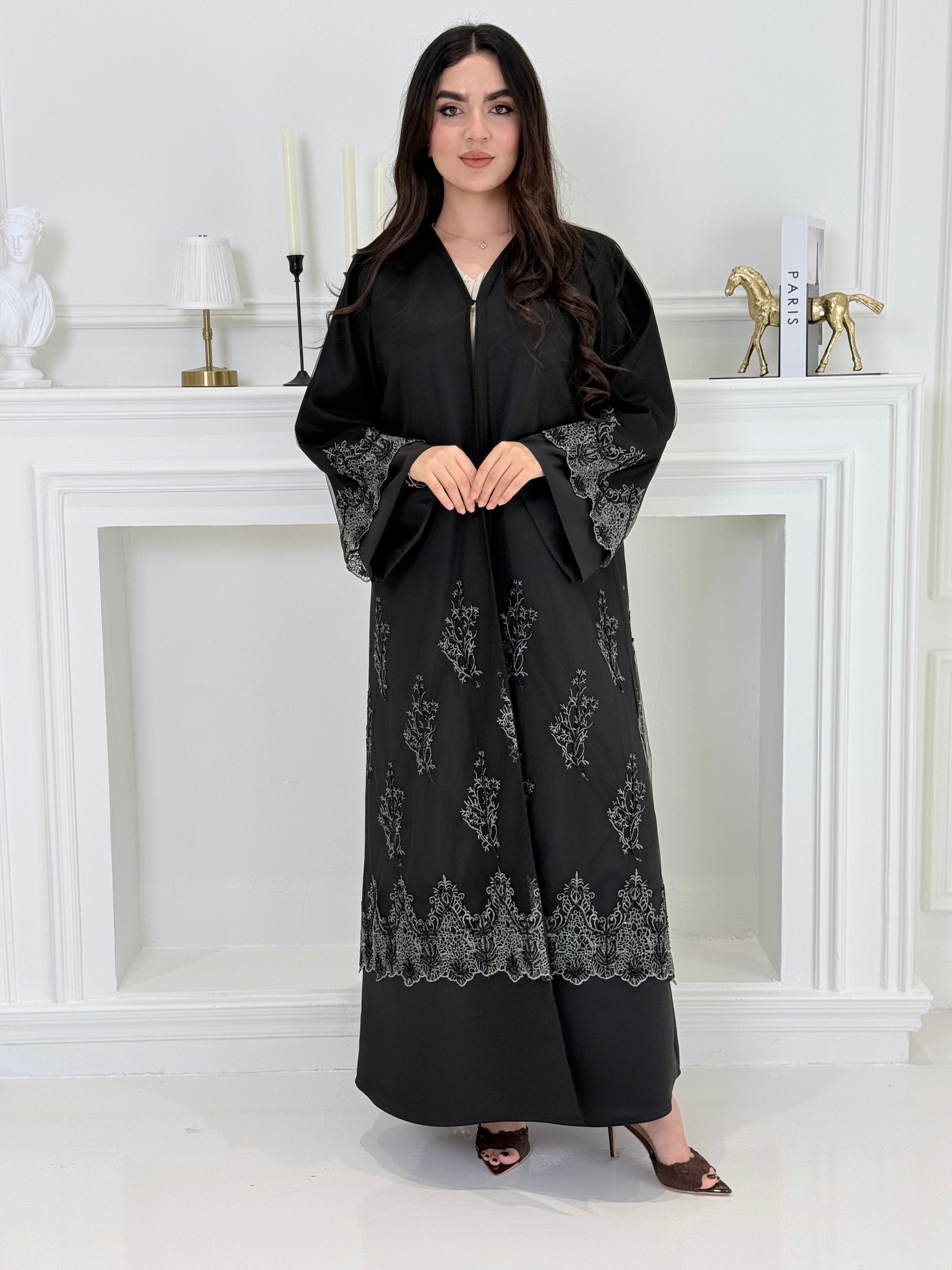 Woman wearing a Premium Silver Embroidered Black Abaya dress with lace details made from Nida fabric and matchhing sheila in a white room.