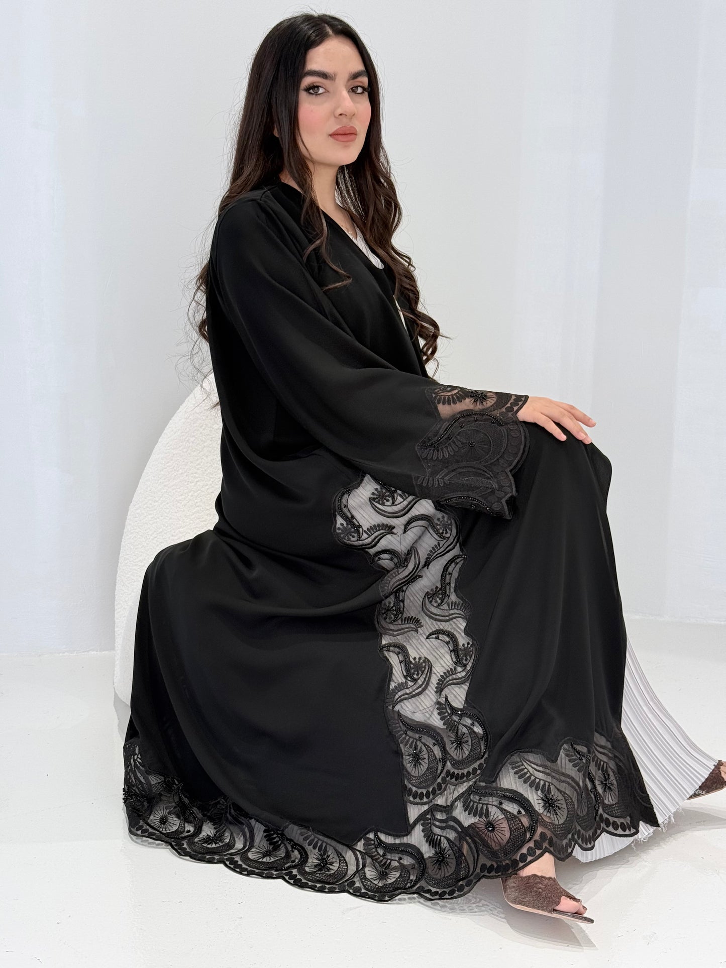 Woman wearing a Premium Black Hand-Embellished Abaya with lace details against a white textured wall.