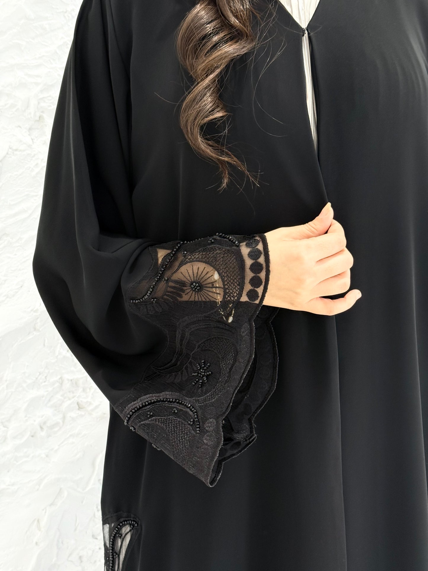 Woman wearing a Premium Black Hand-Embellished Abaya with lace details against a white textured wall.