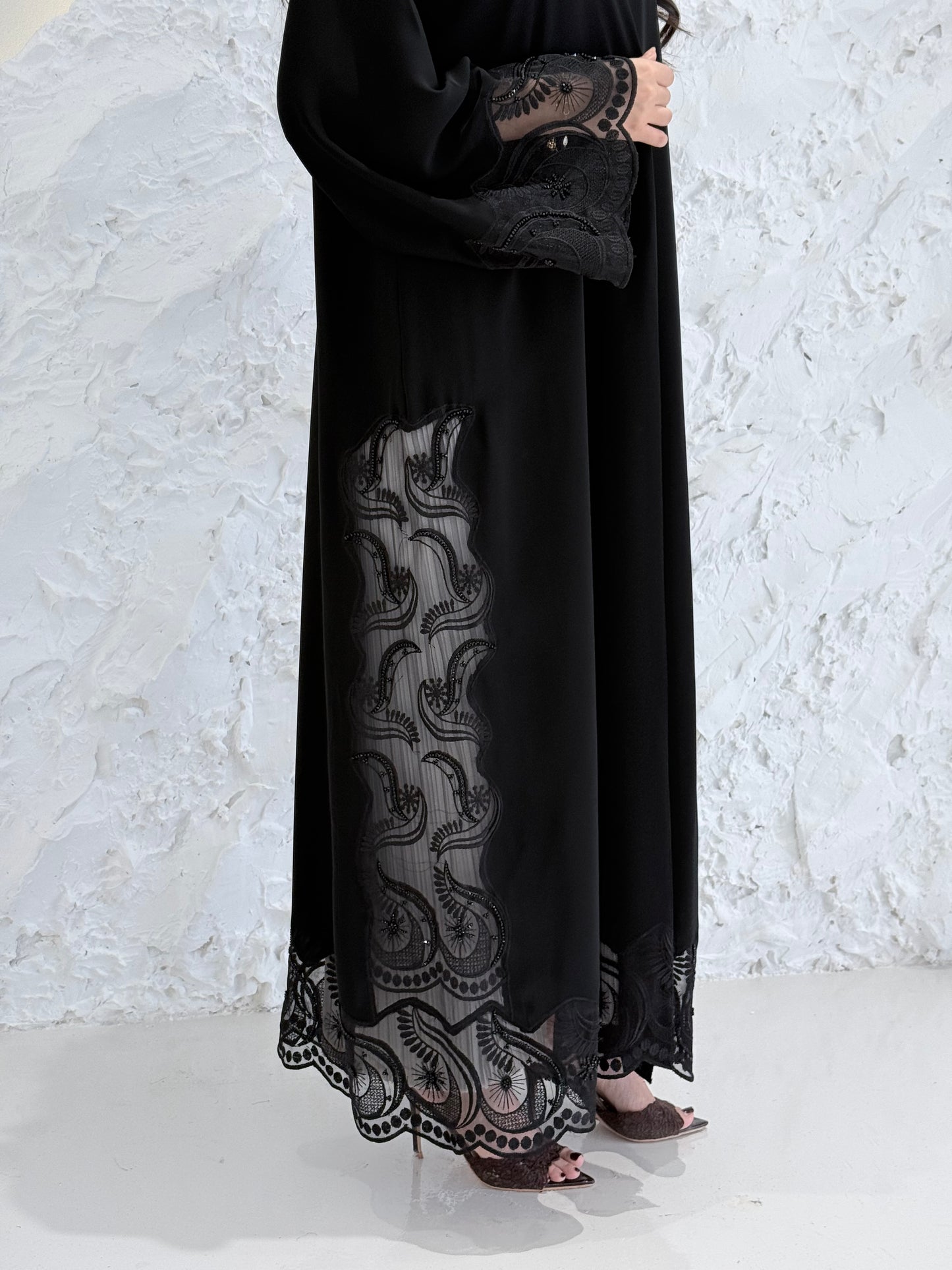 Woman wearing a Premium Black Hand-Embellished Abaya with lace details against a white textured wall.
