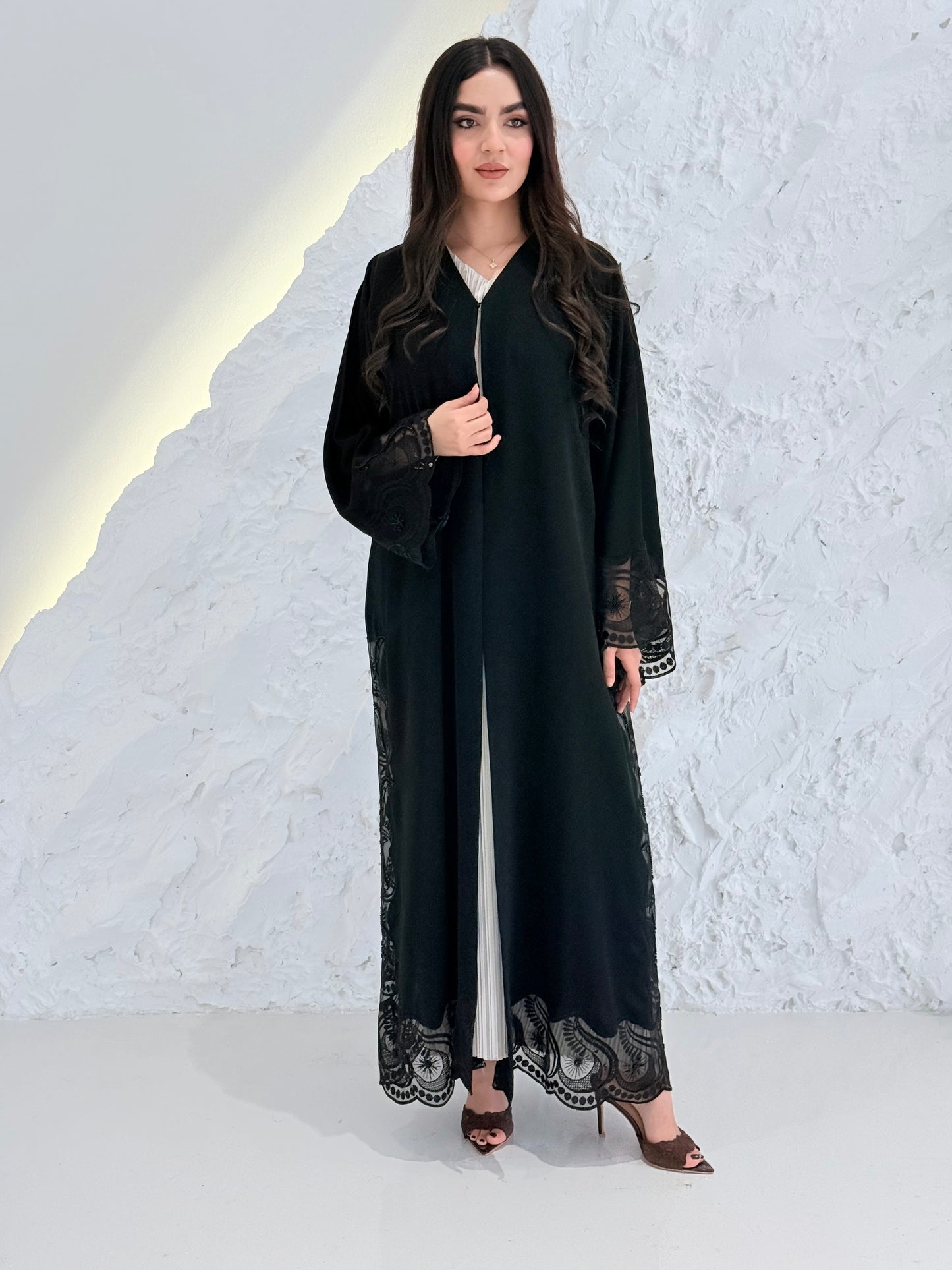 Woman wearing a Premium Black Hand-Embellished Abaya with lace details against a white textured wall.