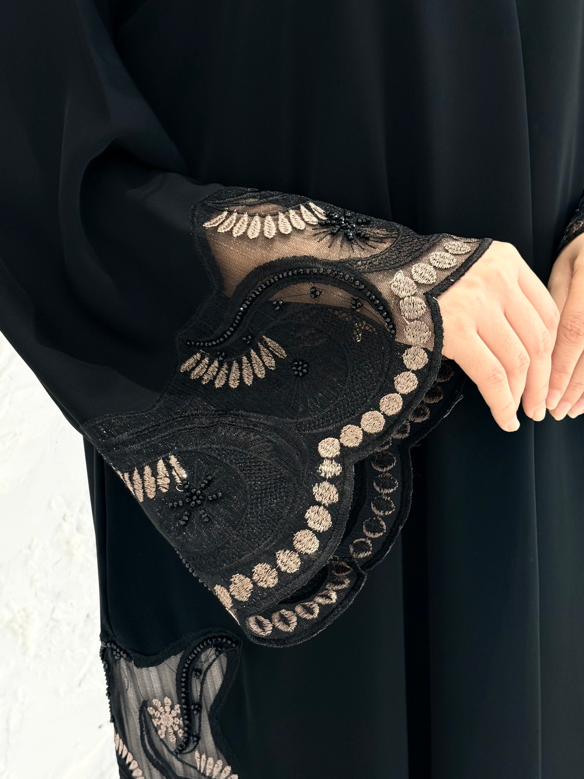 Woman wearing a Premium Gold Hand-Embellished Abaya with lace details against a white textured wall.