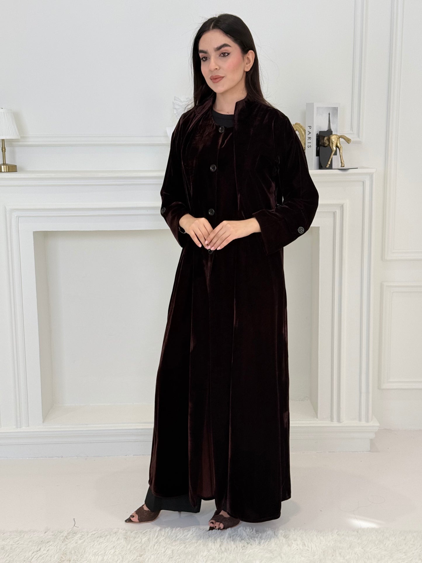 Woman in a long Dark Maroon Velvet Trench Abaya coat standing in a room with white walls and a door.