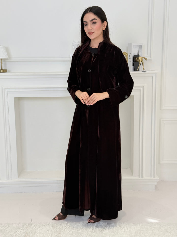 Woman in a long Dark Maroon Velvet Trench Abaya coat standing in a room with white walls and a door.