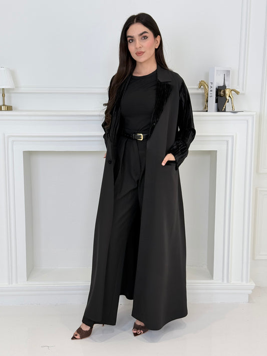 Woman wearing a long black coat-style abaya made from Premium Barbie (Body) and Crushed Velvet in a white interior setting