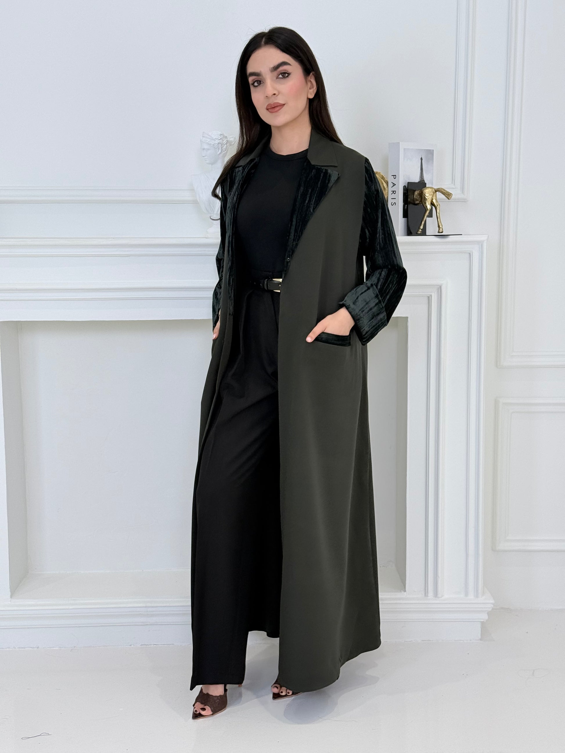 Woman wearing a long olive green coat-style abaya made from Premium Barbie (Body) and Crushed Velvet in a white interior setting