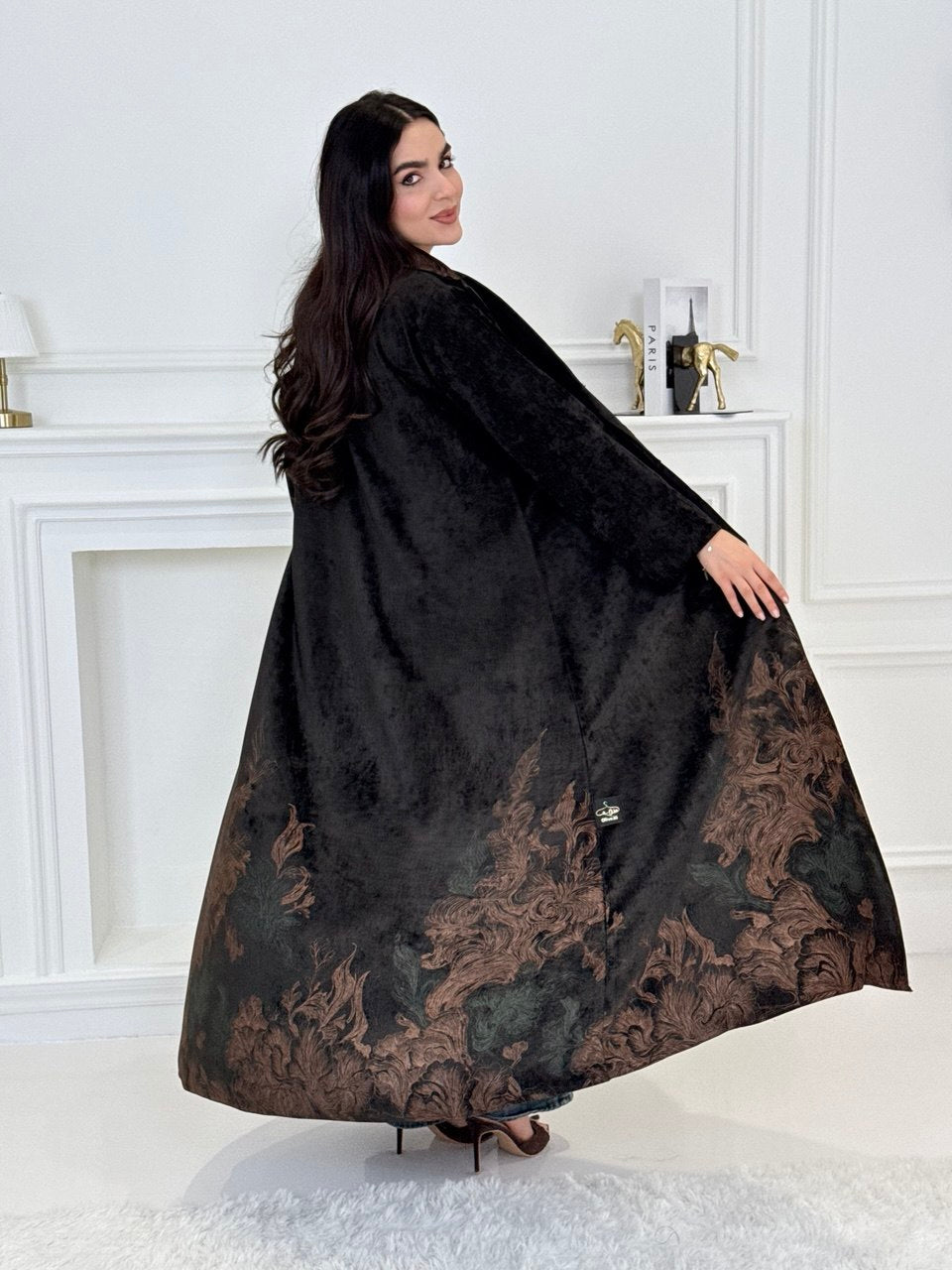 A woman wearing a in Black Velvet Abaya with brown & green Details a white room
