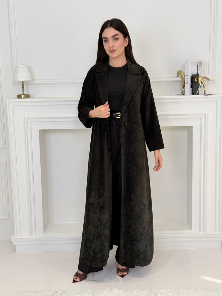 Woman in a long Black Velvet Abaya with Forest Green Details standing in front of a white door.