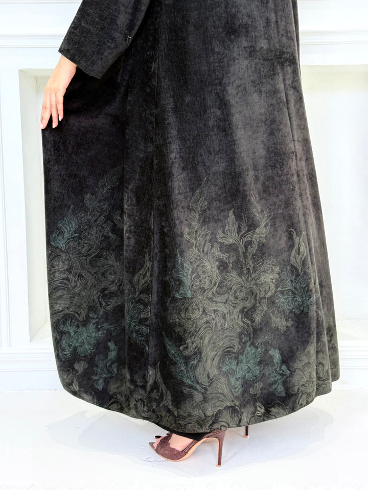 Woman in a long Black Velvet Abaya with Forest Green Details standing in front of a white door.