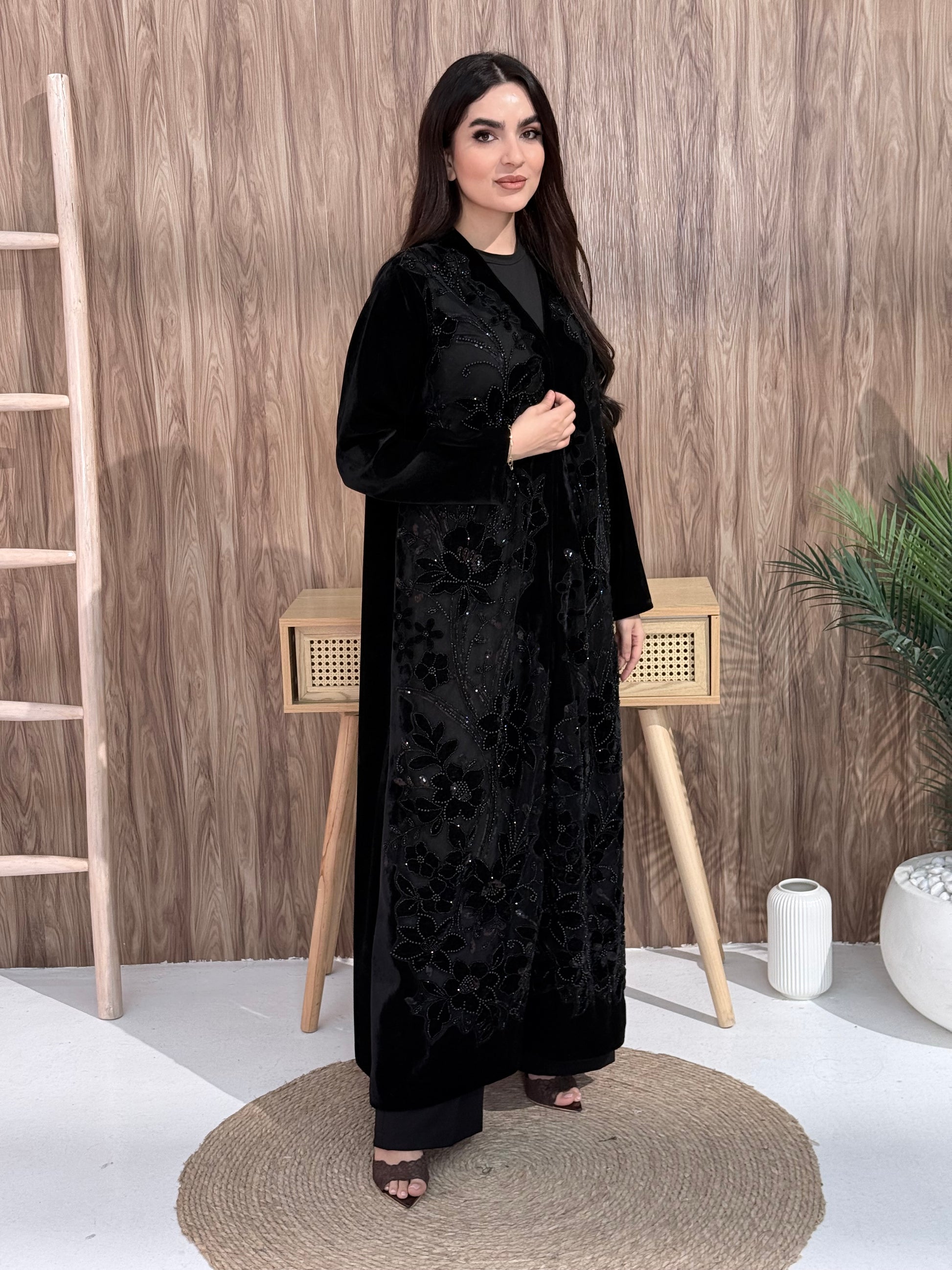 woman wearing a Premium Black Velvet Abaya with Floral Embroidery, lace garment on a wooden floor background
