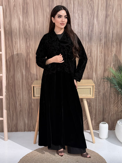 Woman wearing Premium Black Velvet Abaya with Floral Embroidery against a wooden wall.