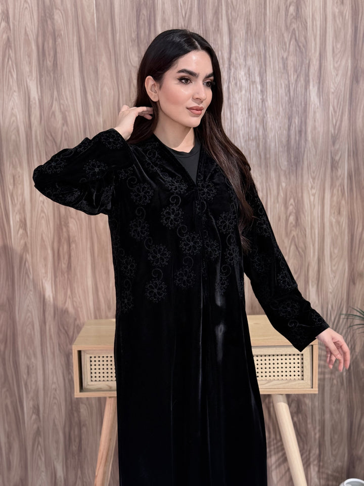 Woman wearing a black velvet coat abaya with intricate patterns against a wooden background