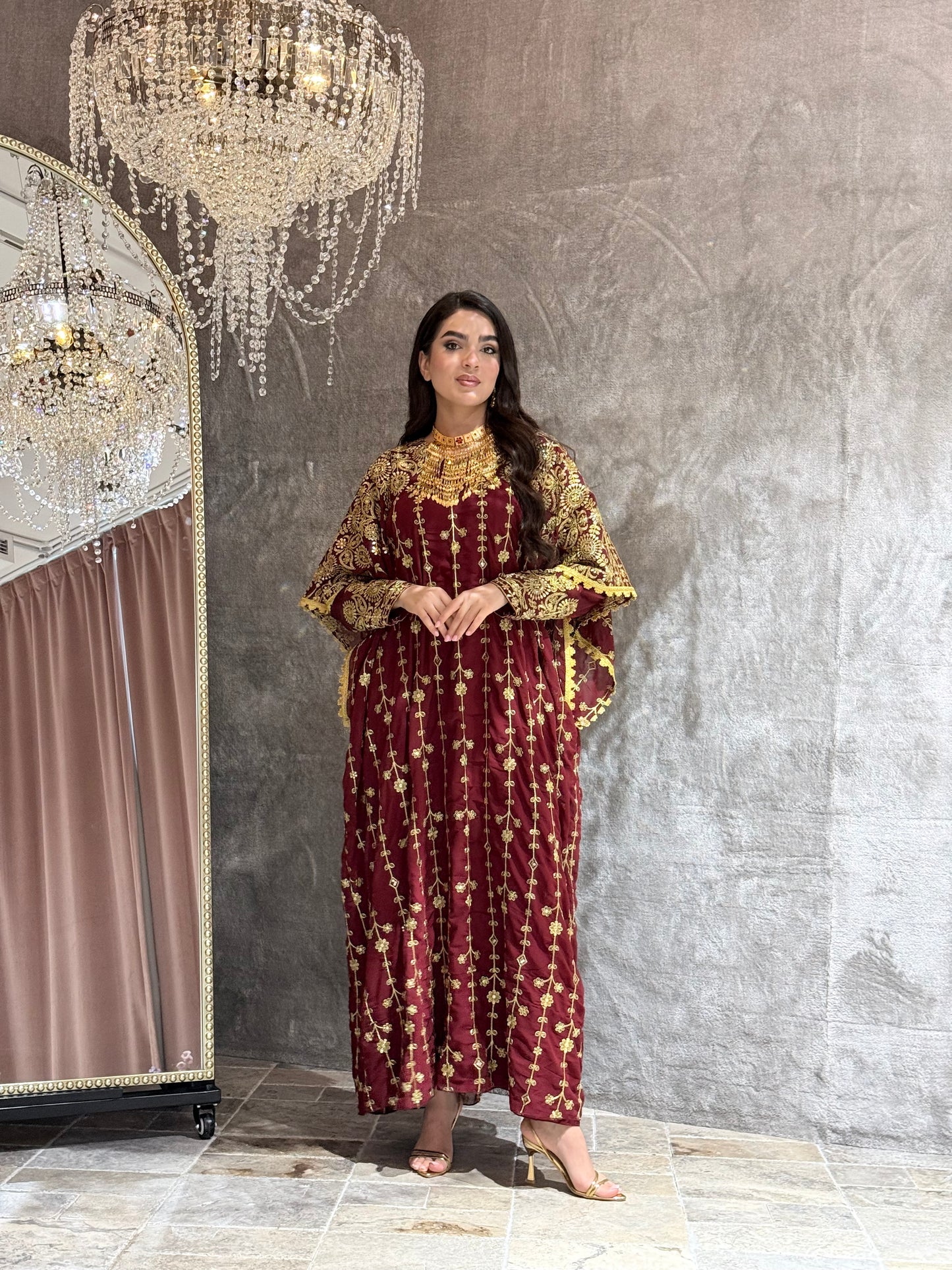 Woman wearing a maroon and gold embroidered kaftan outfit on a textured gray background