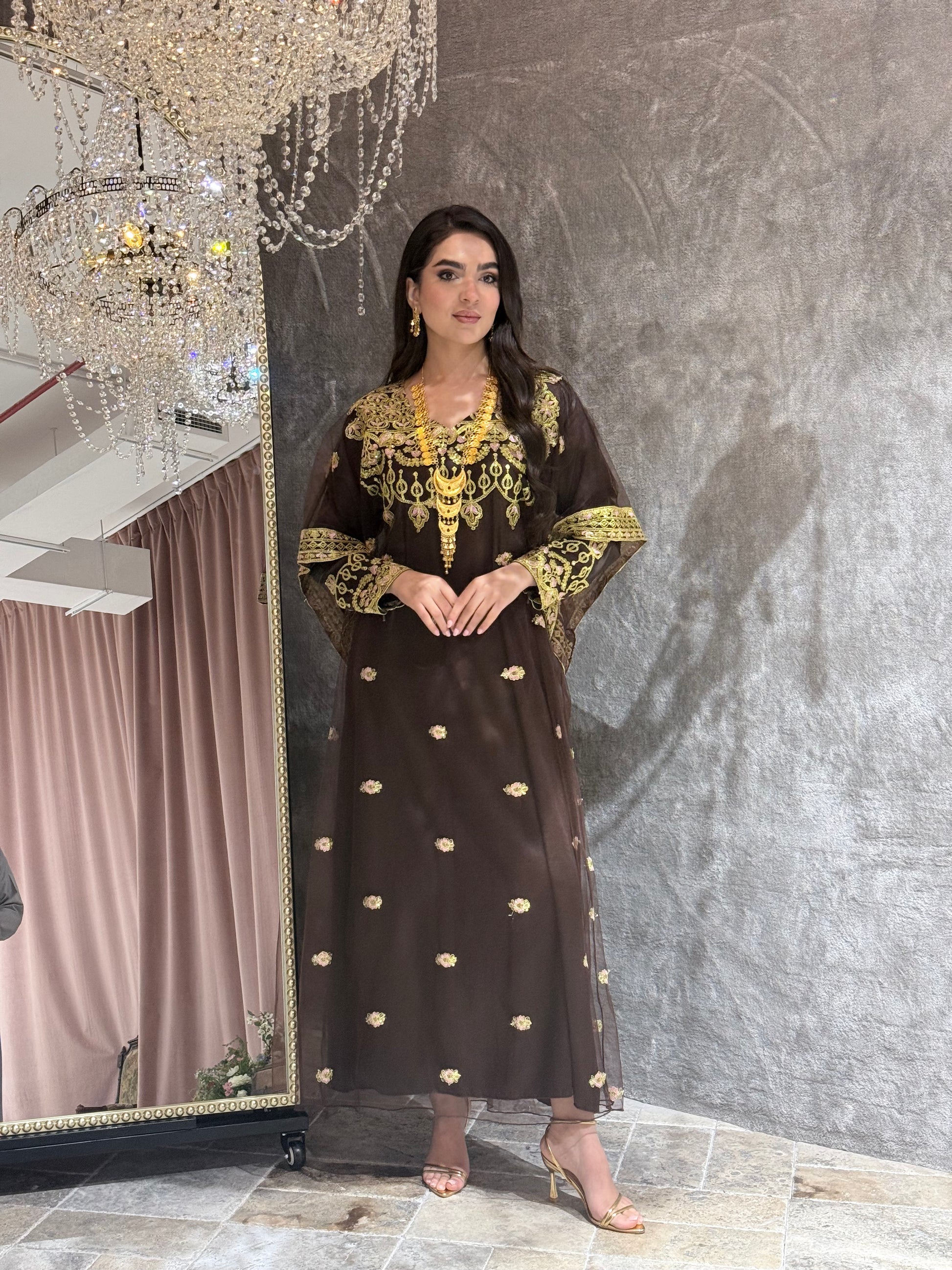 Woman wearing a dark brown traditional kaftan outfit with gold embroidery on a textured gray background