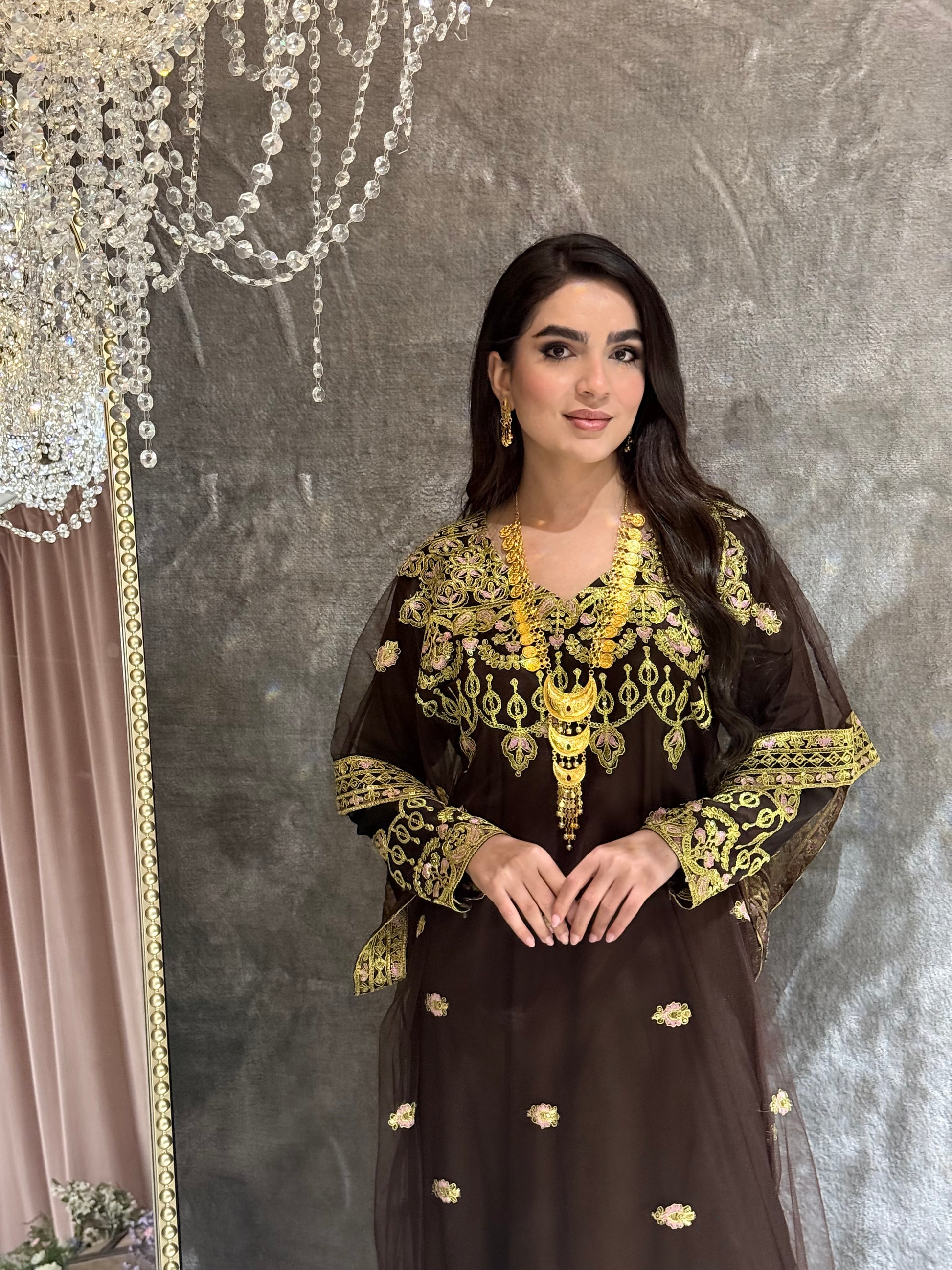 Woman wearing a dark brown traditional kaftan outfit with gold embroidery on a textured gray background