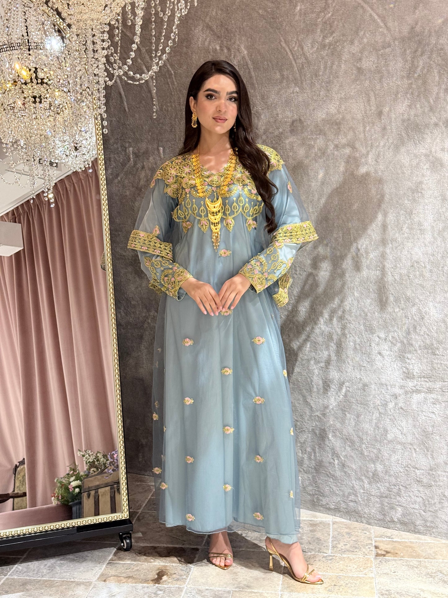Woman in a dusty blue dress with gold Embroidered Kaftan standing in front of a mirror.