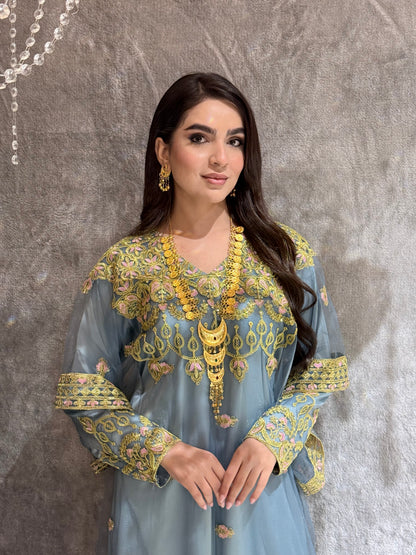 Woman in a dusty blue dress with gold Embroidered Kaftan standing in front of a mirror.