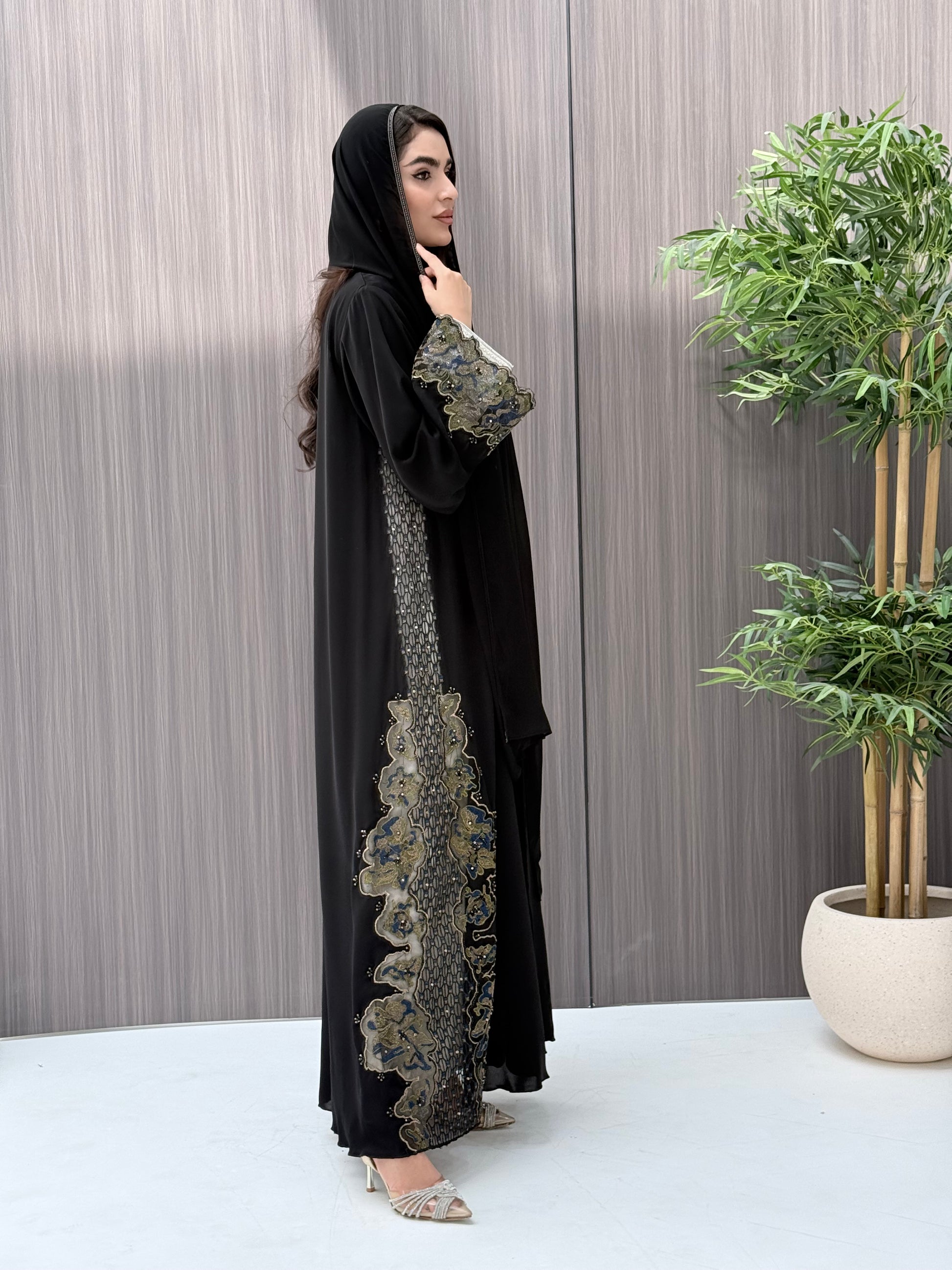 a woman wearing an elegant open-front black abaya featuring intricate multi-colored beadwork, sequins, and metallic embroidery in an indoor setting.