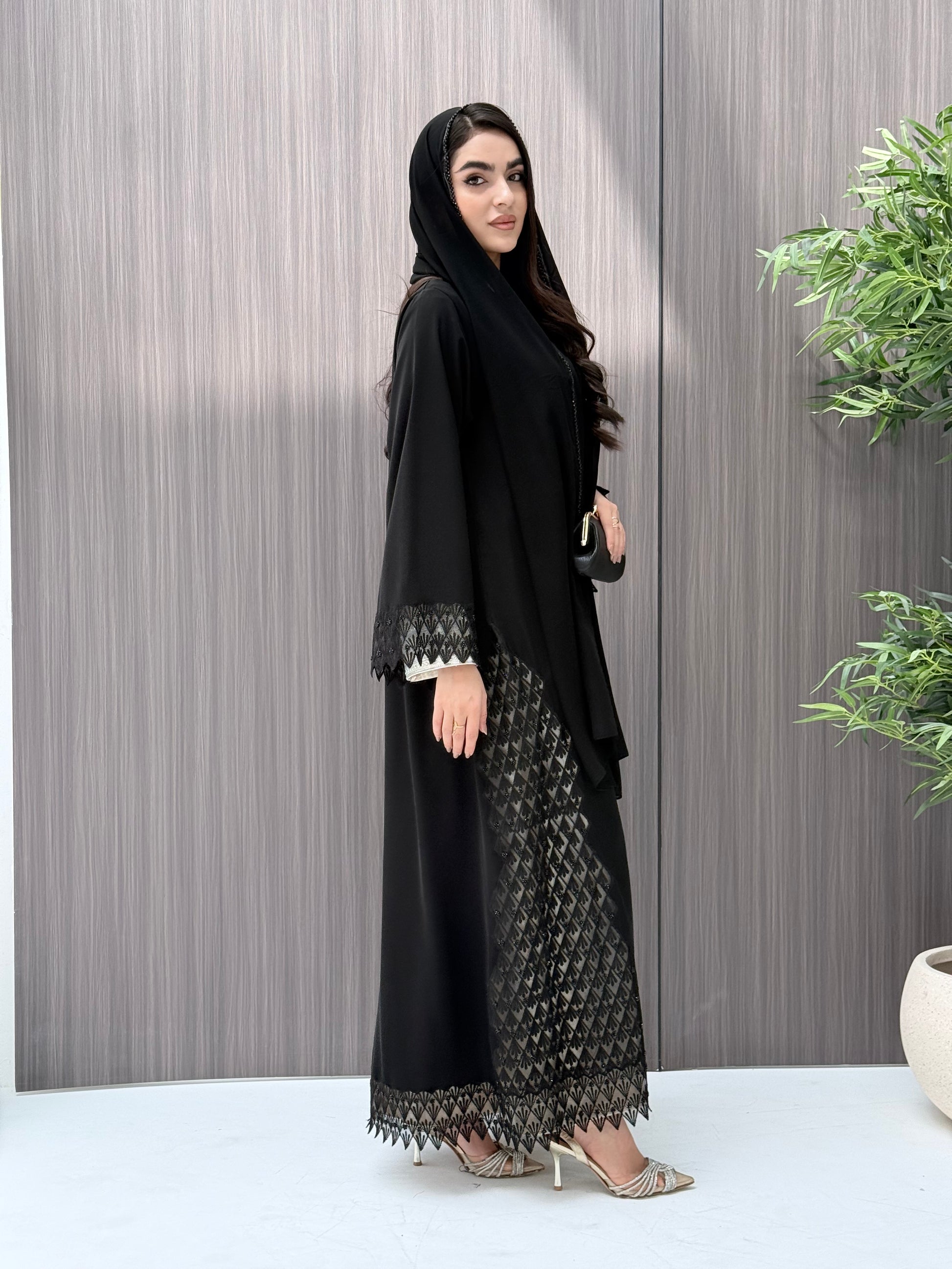a woman wearing a sophisticated luxury open-front black abaya featuring intricate geometric sheer embroidery, elegant lace details, and beaded trims. Includes a matching sheila on a patterned background