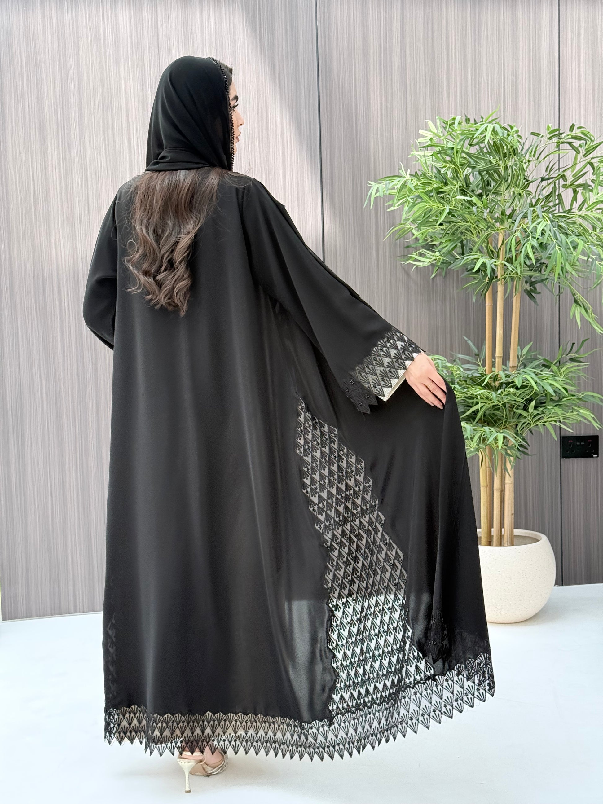 a woman wearing a sophisticated luxury open-front black abaya featuring intricate geometric sheer embroidery, elegant lace details, and beaded trims. Includes a matching sheila on a patterned background