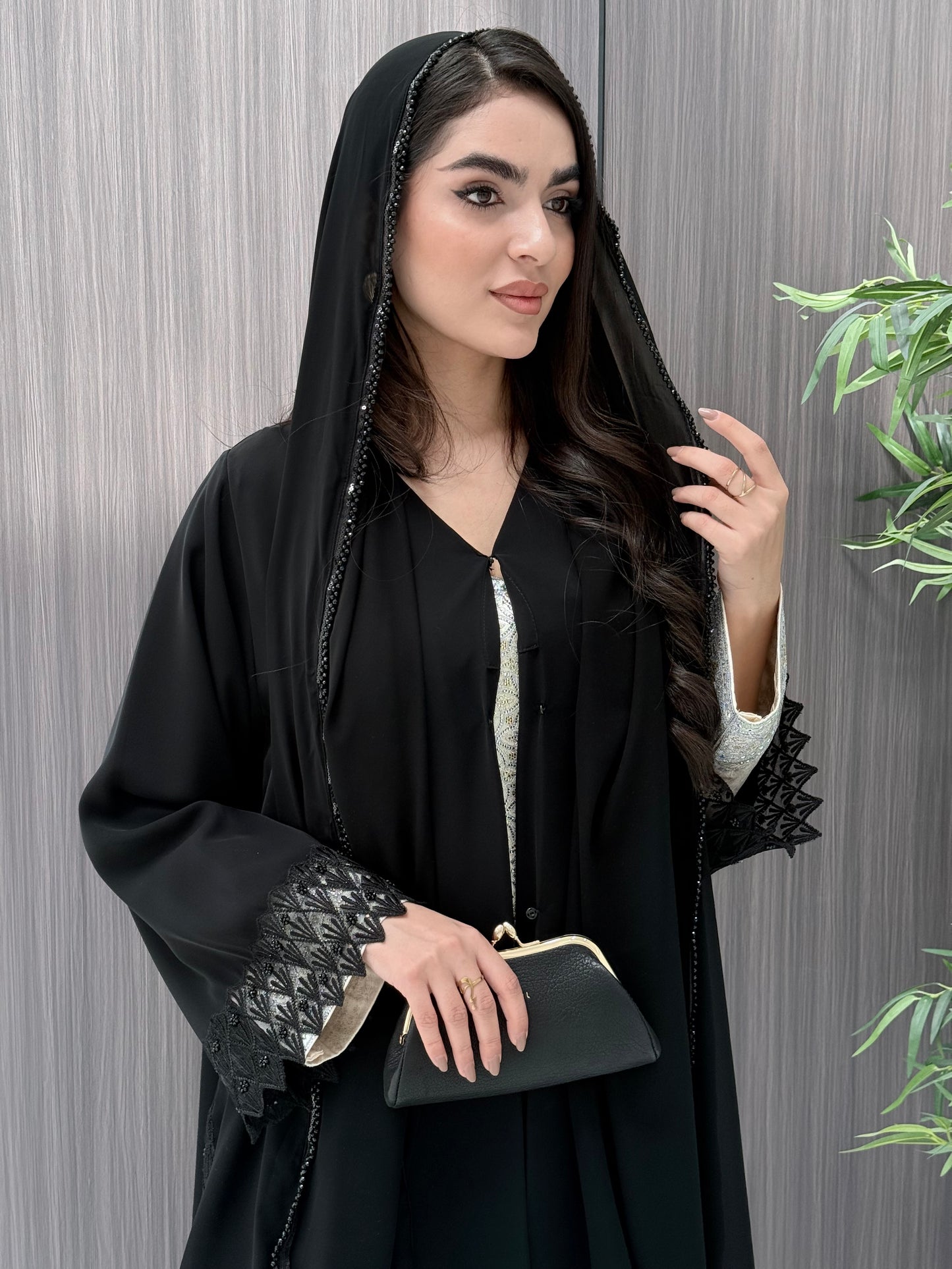 a woman wearing a sophisticated luxury open-front black abaya featuring intricate geometric sheer embroidery, elegant lace details, and beaded trims. Includes a matching sheila on a patterned background