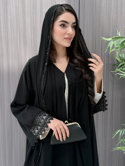 a woman wearing a sophisticated luxury open-front black abaya featuring intricate geometric sheer embroidery, elegant lace details, and beaded trims. Includes a matching sheila on a patterned background