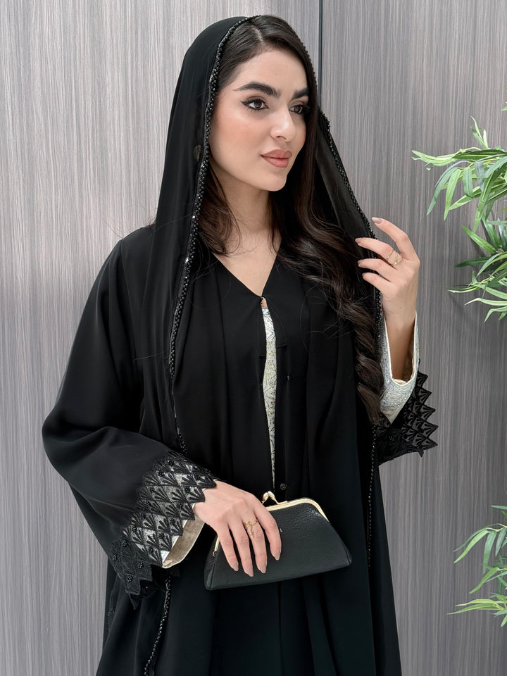 a woman wearing a sophisticated luxury open-front black abaya featuring intricate geometric sheer embroidery, elegant lace details, and beaded trims. Includes a matching sheila on a patterned background