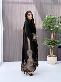 Woman wearing an open-front black abaya featuring intricate sheer panels with bronze, gold, and silver embroidery with matching sheila standing in a room with a plant and lamp.