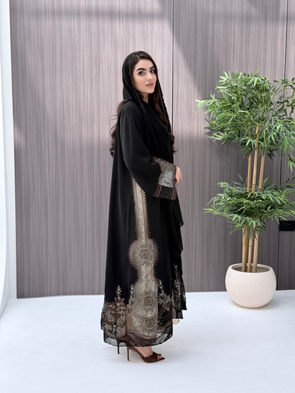 Woman wearing an open-front black abaya featuring intricate sheer panels with bronze, gold, and silver embroidery with matching sheila standing in a room with a plant and lamp.