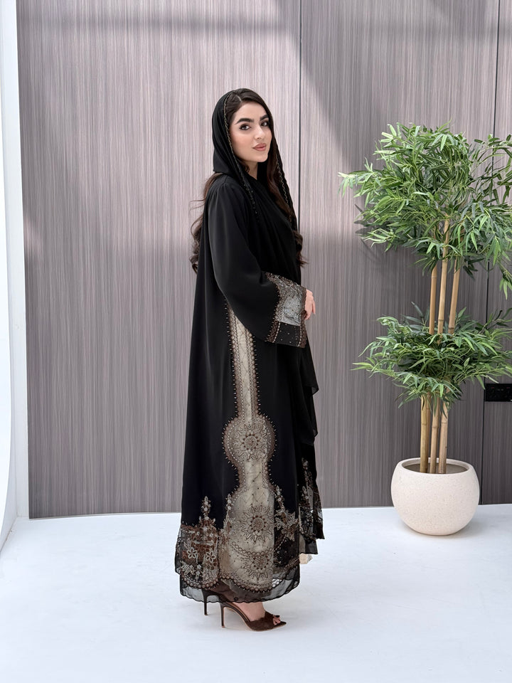 Woman wearing an open-front black abaya featuring intricate sheer panels with bronze, gold, and silver embroidery with matching sheila standing in a room with a plant and lamp.