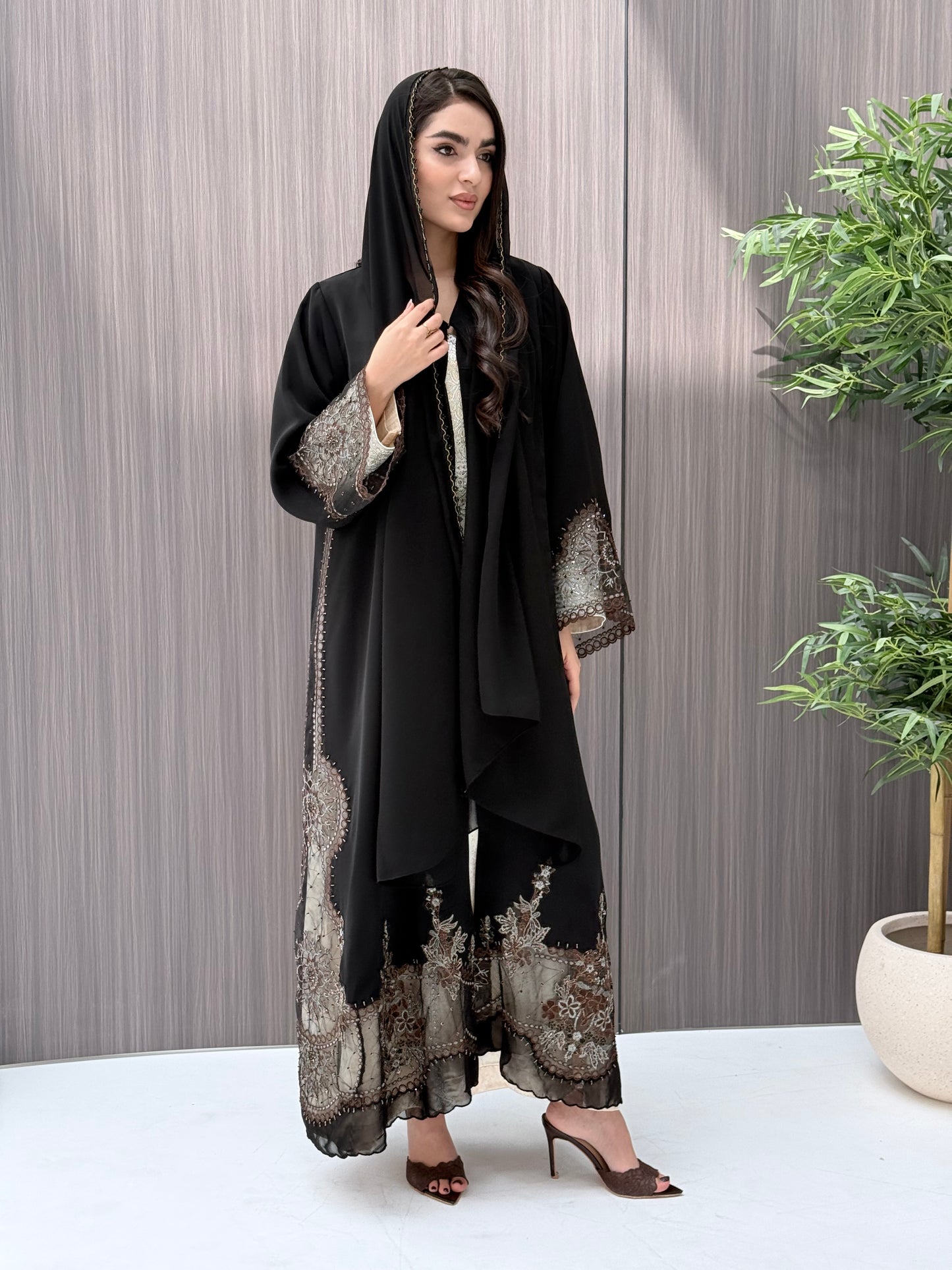 Woman wearing an open-front black abaya featuring intricate sheer panels with bronze, gold, and silver embroidery with matching sheila standing in a room with a plant and lamp.