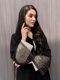 Woman wearing an open-front black abaya featuring intricate sheer panels with bronze, gold, and silver embroidery with matching sheila standing in a room with a brown background