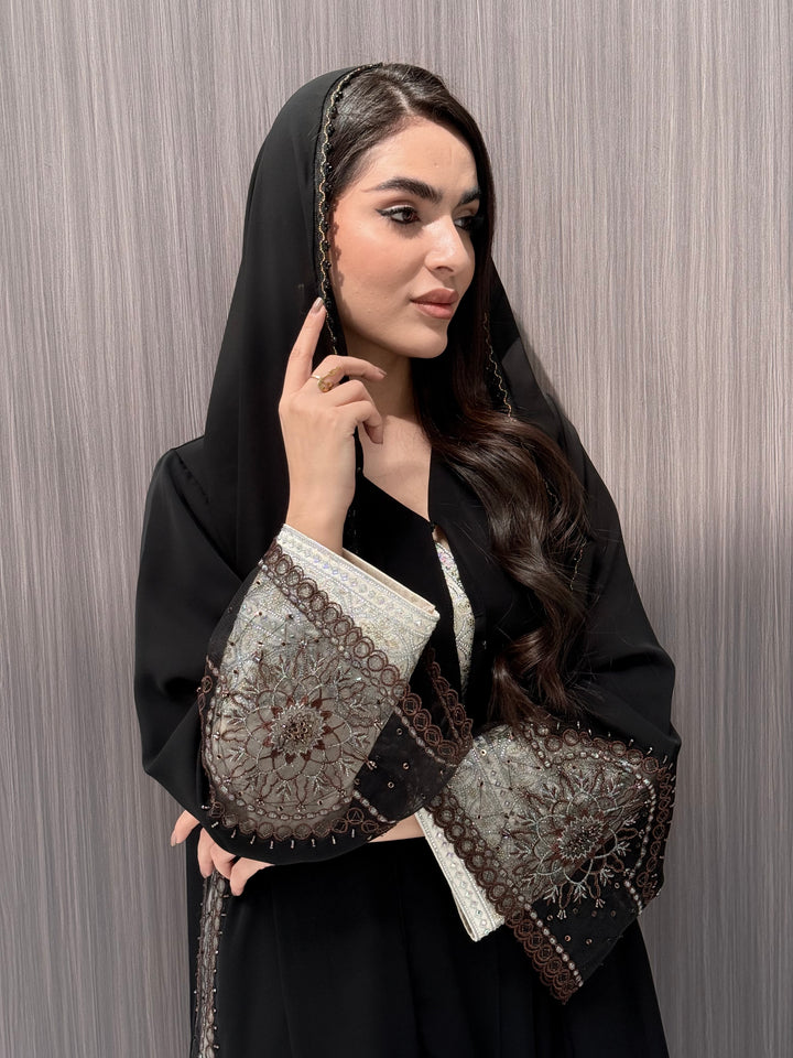 Woman wearing an open-front black abaya featuring intricate sheer panels with bronze, gold, and silver embroidery with matching sheila standing in a room with a brown background