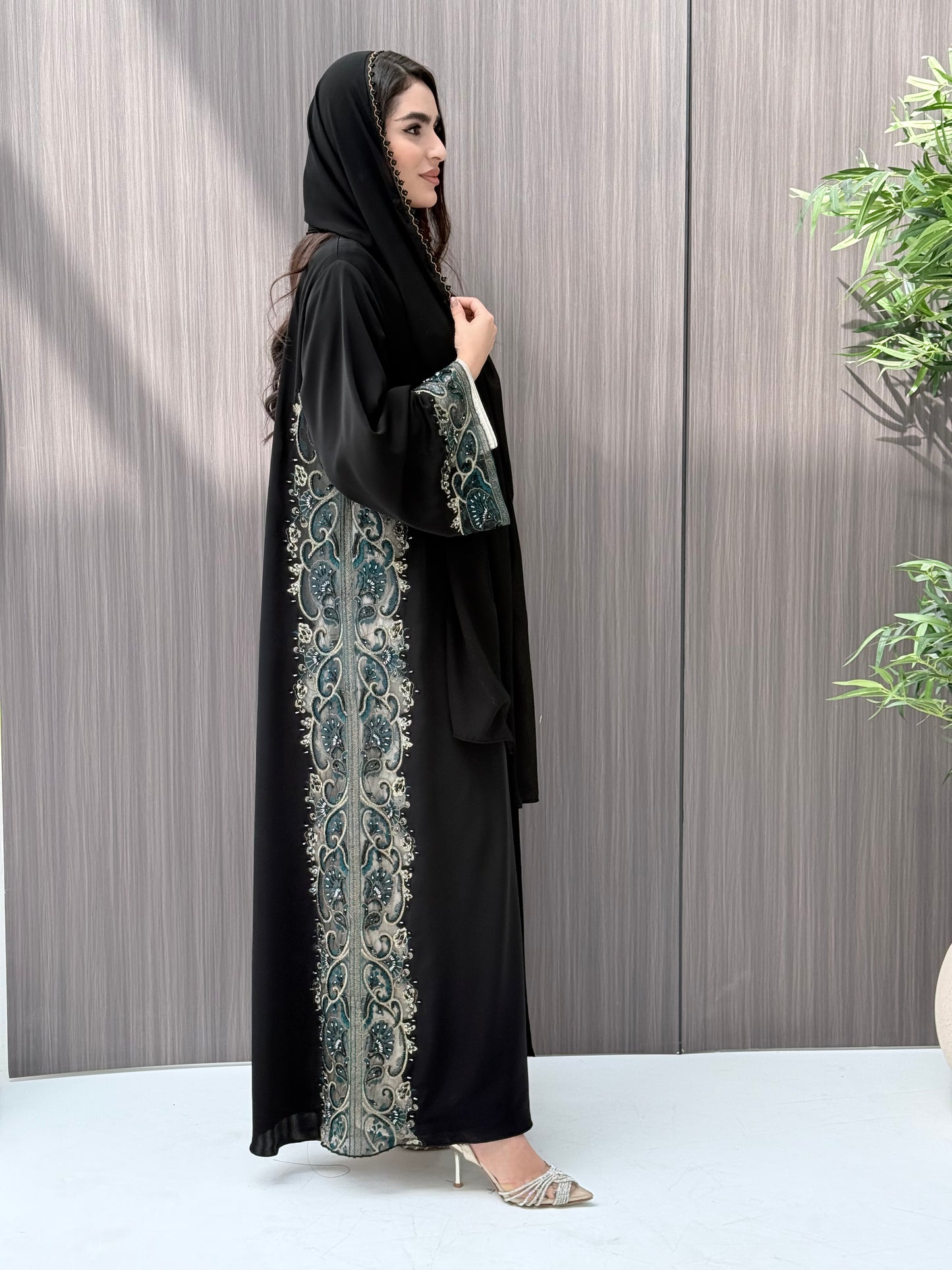 Woman wearing an elegant luxury open-front black abaya crafted from premium Nida fabric, featuring intricate teal, gold, and silver embroidery with delicate beadwork. Includes a perfectly matching sheila on a wooden floor.