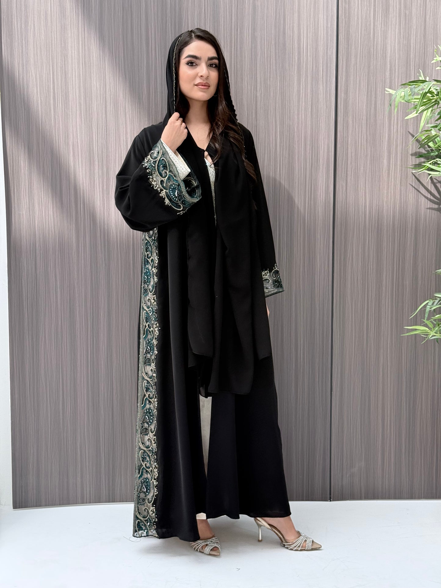 Woman wearing an elegant luxury open-front black abaya crafted from premium Nida fabric, featuring intricate teal, gold, and silver embroidery with delicate beadwork. Includes a perfectly matching sheila on a wooden floor.