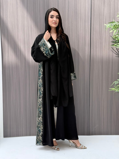 Woman wearing an elegant luxury open-front black abaya crafted from premium Nida fabric, featuring intricate teal, gold, and silver embroidery with delicate beadwork. Includes a perfectly matching sheila on a wooden floor.