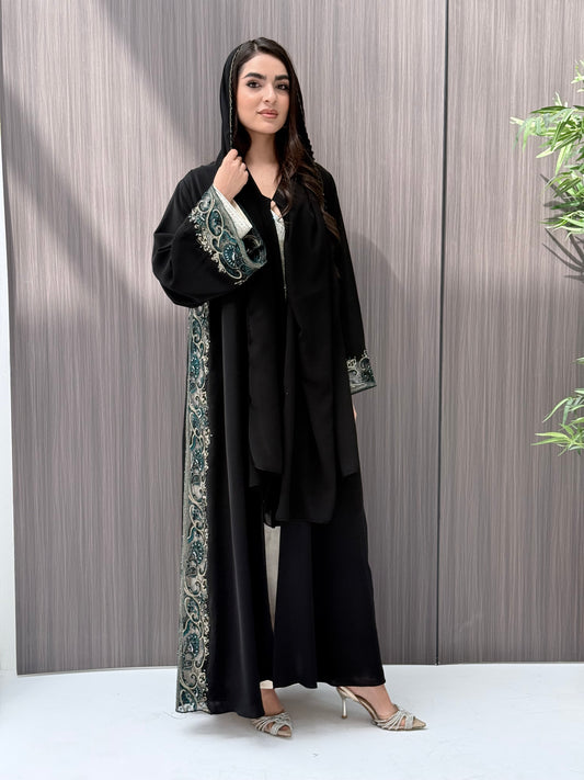 Woman wearing an elegant luxury open-front black abaya crafted from premium Nida fabric, featuring intricate teal, gold, and silver embroidery with delicate beadwork. Includes a perfectly matching sheila on a wooden floor.