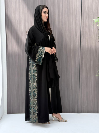 Woman wearing a black abaya with floral patterns standing against a wooden panel background.