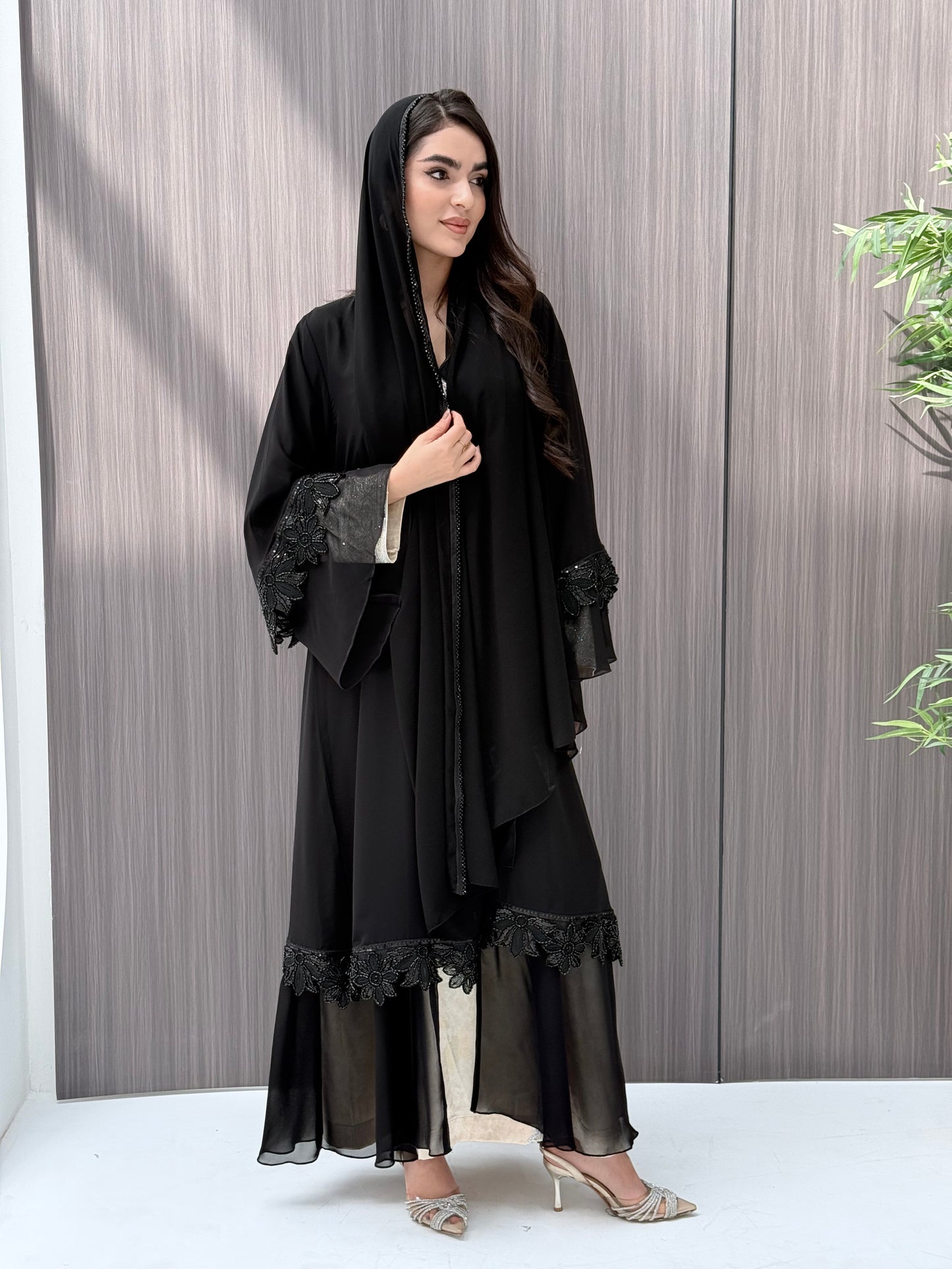 A woman wearing a black beaded, floral open abaya with lace details on a wooden floor.