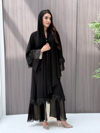 A woman wearing a black beaded, floral open abaya with lace details on a wooden floor.