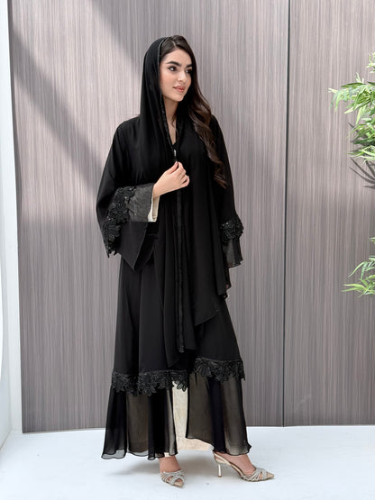 A woman wearing a black beaded, floral open abaya with lace details on a wooden floor.