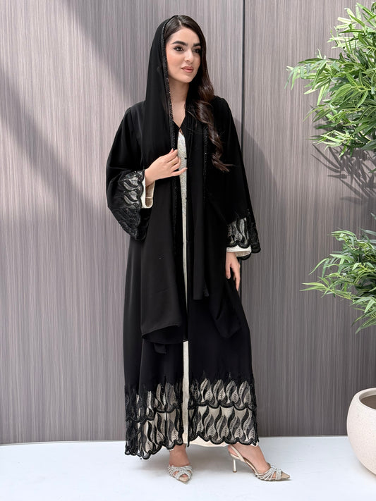 Woman wearing an elegant luxury open-front black abaya crafted from premium Nida fabric, featuring striking sheer panels with botanical leaf embroidery, delicate beadwork, and a matching sheila perfect for Eid, Ramadan, weddings, Henna, parties, and other special occasions.
 in a minimal indoor setting
