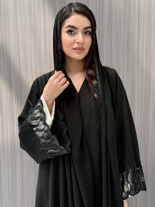 Woman wearing an elegant luxury open-front black abaya crafted from premium Nida fabric, featuring striking sheer panels with botanical leaf embroidery, delicate beadwork, and a matching sheila perfect for Eid, Ramadan, weddings, Henna, parties, and other special occasions.
 in a minimal indoor setting