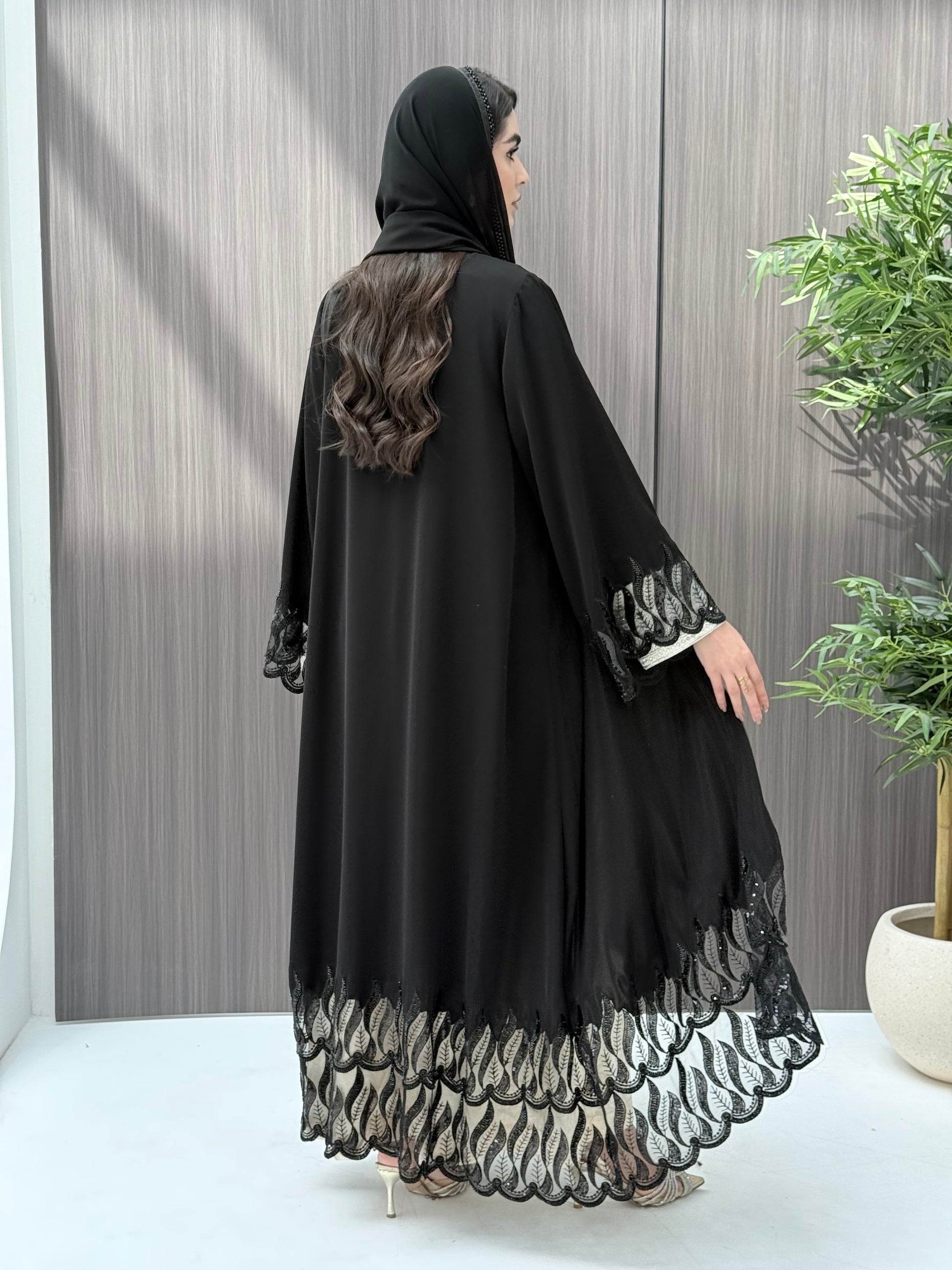 Woman wearing an elegant luxury open-front black abaya crafted from premium Nida fabric, featuring striking sheer panels with botanical leaf embroidery, delicate beadwork, and a matching sheila perfect for Eid, Ramadan, weddings, Henna, parties, and other special occasions.
 in a minimal indoor setting