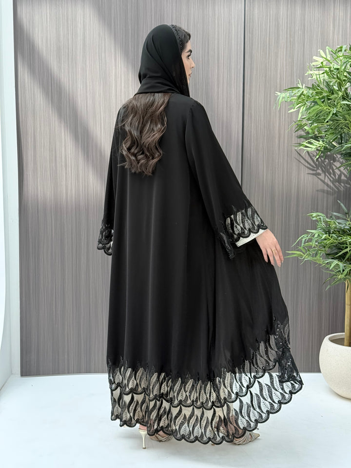 Woman wearing an elegant luxury open-front black abaya crafted from premium Nida fabric, featuring striking sheer panels with botanical leaf embroidery, delicate beadwork, and a matching sheila perfect for Eid, Ramadan, weddings, Henna, parties, and other special occasions.
 in a minimal indoor setting
