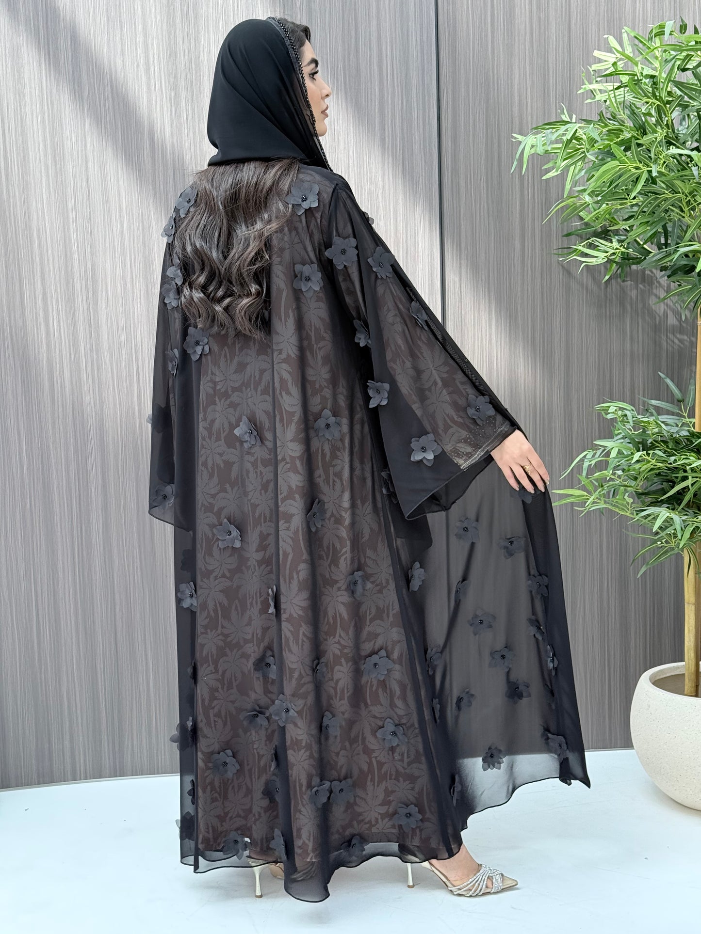 Woman wearing an open-front sheer black abaya featuring elegant 3D floral appliqués. Complete with a matching beaded sheila, perfect for weddings, Eid, and special occasions standing on a wooden floor.