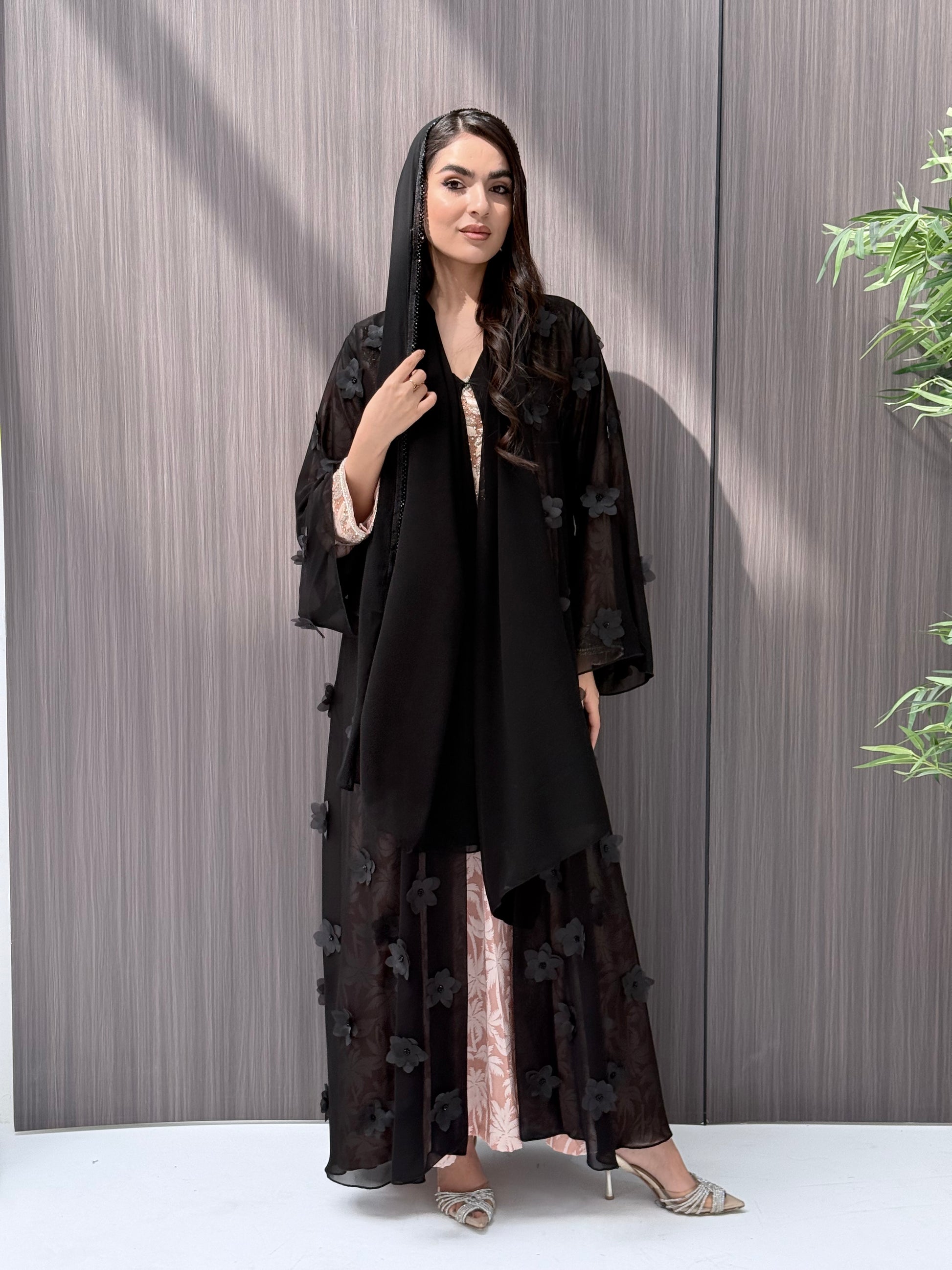 Woman wearing an open-front sheer black abaya featuring elegant 3D floral appliqués. Complete with a matching beaded sheila, perfect for weddings, Eid, and special occasions standing on a wooden floor.