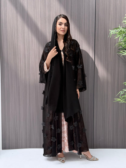 Woman wearing an open-front sheer black abaya featuring elegant 3D floral appliqués. Complete with a matching beaded sheila, perfect for weddings, Eid, and special occasions standing on a wooden floor.