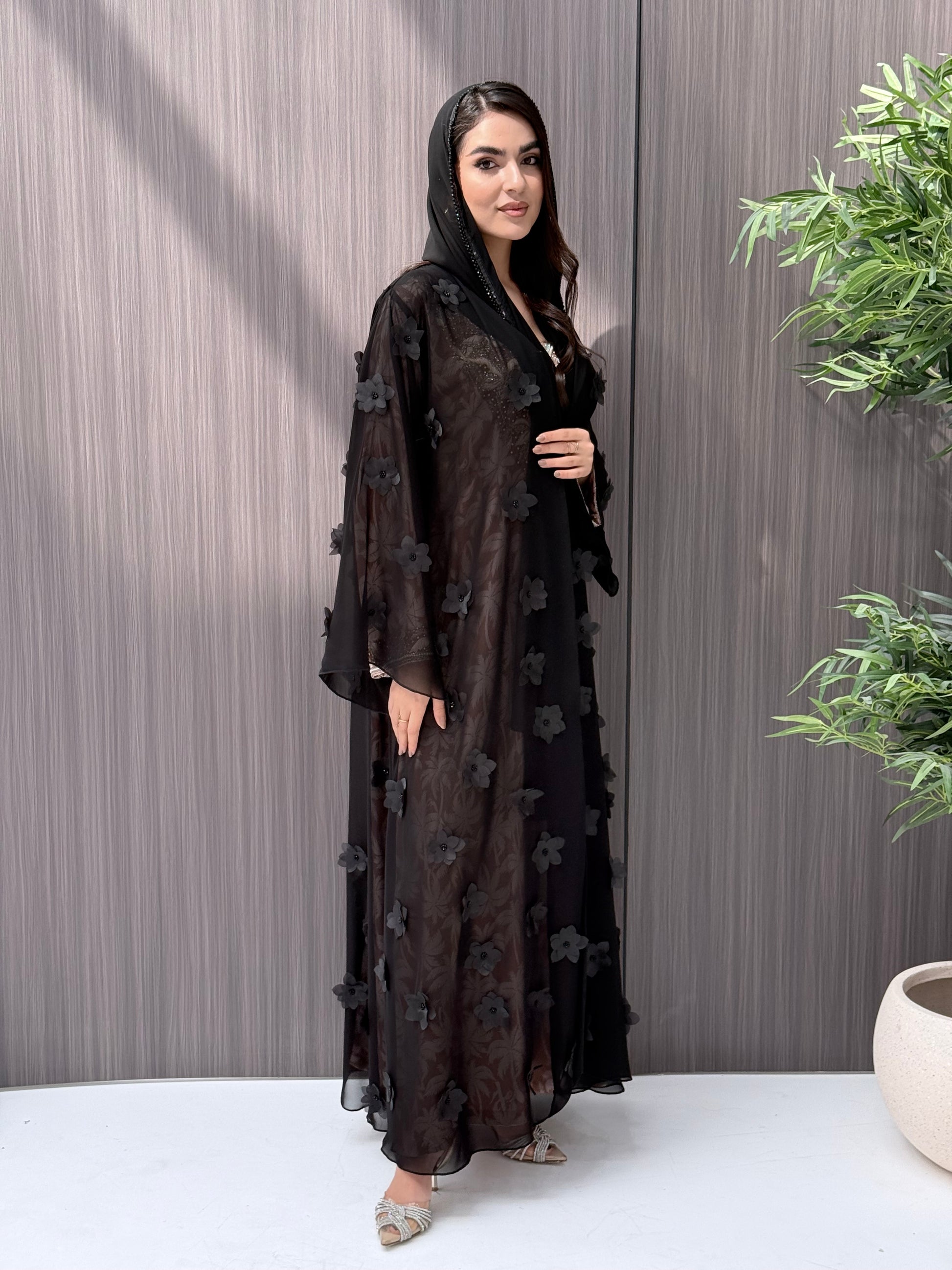 Woman wearing an open-front sheer black abaya featuring elegant 3D floral appliqués. Complete with a matching beaded sheila, perfect for weddings, Eid, and special occasions standing on a wooden floor.