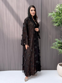 Woman wearing an open-front sheer black abaya featuring elegant 3D floral appliqués. Complete with a matching beaded sheila, perfect for weddings, Eid, and special occasions standing on a wooden floor.